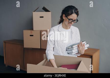 Die junge Frau packt Bücher in den Karton, während sie im Apartment-Zimmer umzieht Stockfoto