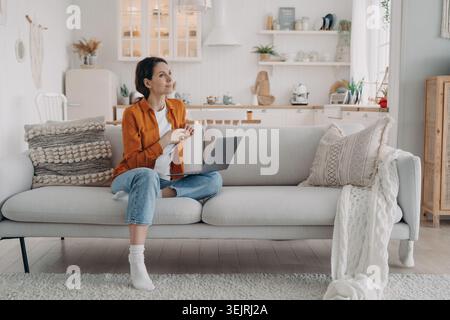 Frau trinkt Kaffee, während sie einen Laptop auf dem Sofa in der hellen Wohnküche benutzt Stockfoto