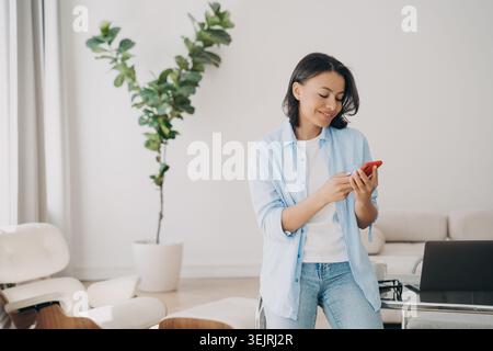 Die junge Frau sucht im hellen, modernen Heimbüro des Wohnzimmers neben dem Laptop nach dem Smartphone Stockfoto