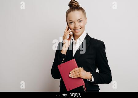 Lächelnde junge Geschäftsfrau in schwarzem Blazer, die auf dem Smartphone spricht, während sie den roten Planer vor neutralem Studiohintergrund hält Stockfoto