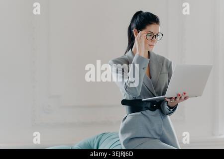 Zielgerichtete Geschäftsfrau in grauem Hosenanzug und Brille hält den Laptop offen, während sie im minimalistischen weißen Innenraum mit Kopierspac liest Stockfoto