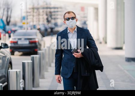 Geschäftsmann mit Gesichtsmaske und Sonnenbrille läuft mit Smartphone auf dem Bürgersteig der Stadt Stockfoto
