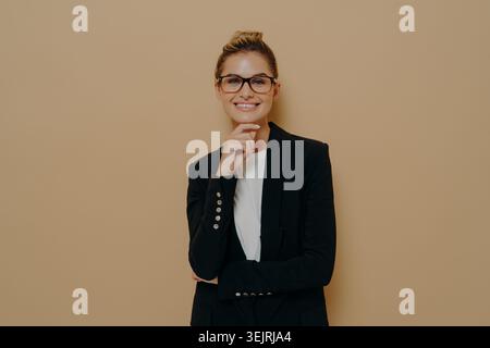Die junge Geschäftsfrau in Brille lächelt mit Hand am Kinn vor der beigefarbenen Atelierwand Stockfoto
