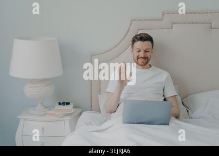 Junger Mann winkt während eines Videogesprächs auf dem Laptop, während er zu Hause im Bett sitzt Stockfoto