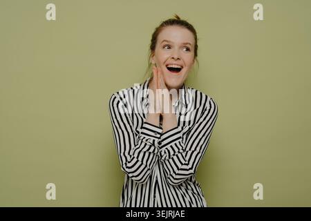 Aufgeregte junge Frau in gestreiftem Hemd schließt die Hände, während sie gegen die grüne Studiowand blickt Stockfoto
