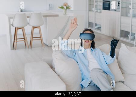Eine junge Frau mit Virtual-Reality-Headset und Roboterhandschuh interagiert mit digitalen Inhalten in einem modernen Zuhause Stockfoto