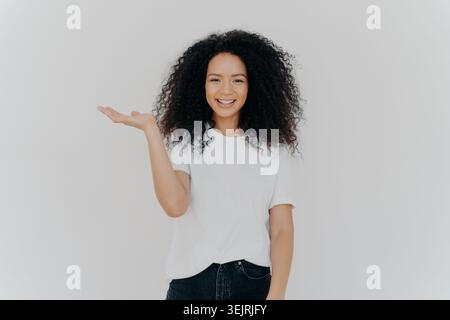 Titel: Lächelnde junge Frau mit lockigen Haaren zeigt offene Hand in Richtung Kopierraum im minimalen Studio Stockfoto