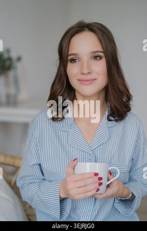 Junge brünette Frau im gestreiften Pyjama, die an einem ruhigen Morgen zu Hause einen weißen Becher hält Stockfoto