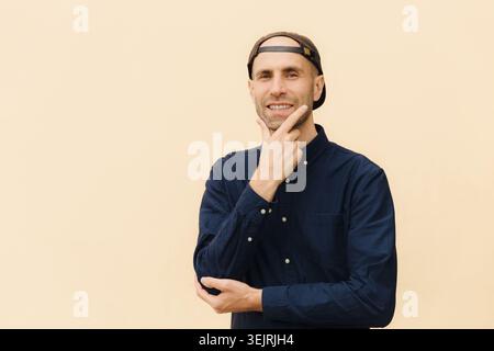 Ein lächelnder, lässiger Mann in einer rückwärtigen Baseballmütze berührt das Kinn an der beigefarbenen Wand Stockfoto
