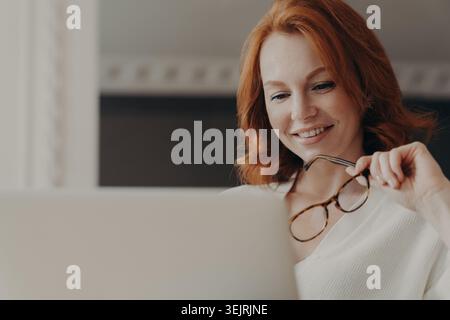 Die rothaarige Frau lächelt, während sie am Laptop arbeitet und zu Hause eine Brille hält Stockfoto
