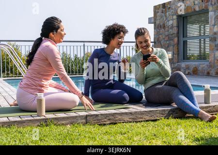 Verschiedene weibliche Freunde in bunten Tops sitzen auf Deck am Pool, teilen sich Telefon und Wasserflaschen. Garten, Outdoor, Yoga, Fitne Stockfoto