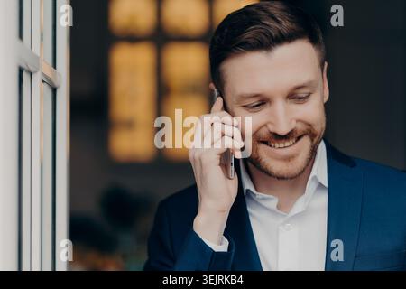 Geschäftsmann lächelt, während er auf dem Smartphone in der Nähe des Bürofensters spricht Stockfoto