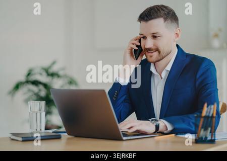 Geschäftsmann spricht über Smartphone, während er am Schreibtisch am Laptop arbeitet Stockfoto