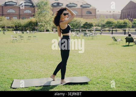 FIT Junge Frau in Schwarz Sportswear steht auf Yogamatte auf grünem Rasen im Urban Park und blickt am sonnigen Tag über die Schulter zurück Stockfoto