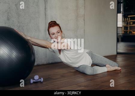 Junge Rothaarige Frau, die auf dem Fitnessraum neben dem Fitnessball liegt und die Hantel lächelt während der Trainingspause im Fitnessstudio Stockfoto