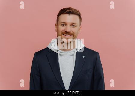 Der lächelnde Kaukasier trägt einen Blazer über dem Hoodie vor rosa Studiohintergrund Stockfoto