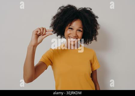 Junge Schwarze Frau lächelt, während sie kleine Gesten mit Fingern in minimalem Studio zeigt Stockfoto