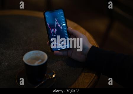 Hand hält Smartphone mit Finanzhandelskarte am dunklen Café-Tisch Stockfoto