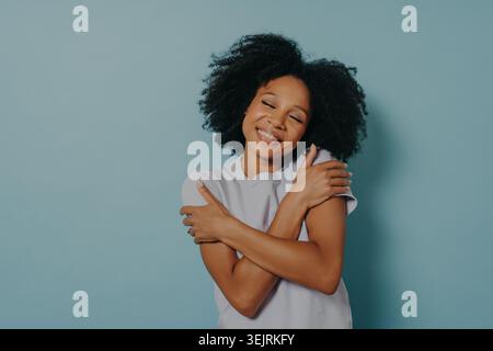 Die junge schwarze Frau lächelt, während sie sich an die blaue Studiowand umarmt Stockfoto
