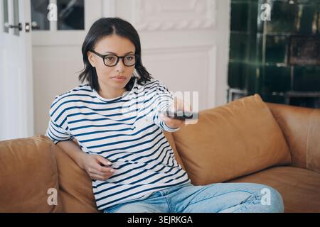 Eine junge Frau mit Brille benutzt die Fernbedienung, während sie sich auf dem Ledersofa im Wohnzimmer entspannt Stockfoto