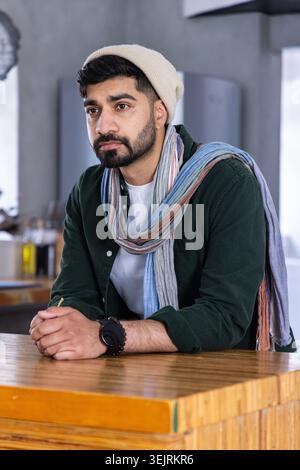 Mann lehnt sich auf Holztheke in einem modernen Café, trägt Mütze und langen mehrfarbigen Schal. Bart, Hut, Uhr, Armband, Porträts Stockfoto