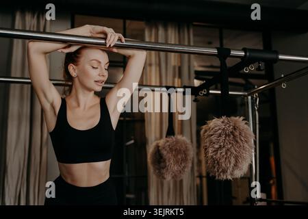 Junge Frau in schwarzer Sportswear, die sich auf Pilates-Reformer-Geräten im Fitness-Studio ausdehnt Stockfoto