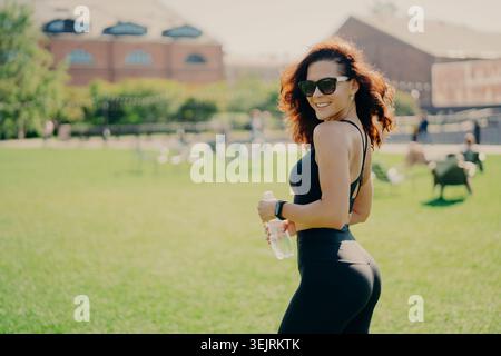 Sportliche Frau in schwarzer Sportbekleidung hält eine Wasserflasche und lächelt im sonnigen Stadtpark Stockfoto