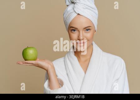 Junge Frau in weißem Bademantel und Handtuch hält grünen Apfel für Beauty- und Wellness-Porträt Stockfoto
