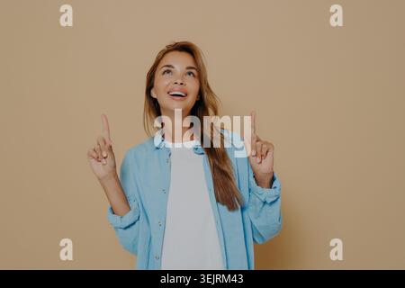 Junge Frau in blauem Denim-Hemd, die beide Zeigefinger nach oben zeigt und mit Begeisterung gegen Beige Studio Backgroun aufblickt Stockfoto