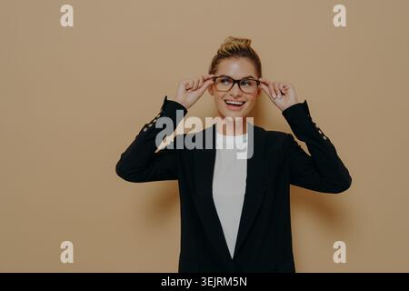 Die junge Geschäftsfrau passt die Brille an und lächelt in schwarzem Blazer gegen die beige Studiowand Stockfoto