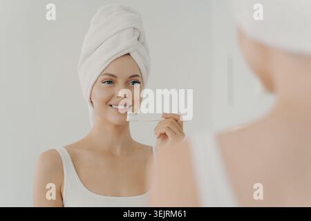 Junge Frau putzt die Zähne mit einer weißen Zahnbürste, während sie im hellen Badezimmer ein Handtuch auf dem Kopf trägt Stockfoto
