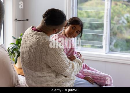 Vater hält ein Mädchen, das auf dem Sofa am offenen Fenster sitzt, und trägt einen cremefarbenen, rosa gestreiften Pyjama Stockfoto
