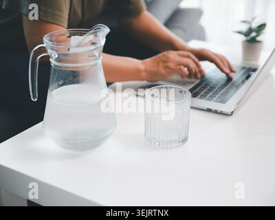 Wasserkrug aus Glas und Becher auf weißem Schreibtisch mit verschwommener Person, die auf Laptop schreibt, zeigt Hydration, Wellness, Fernarbeit, gesunde Büroroutine, Stockfoto