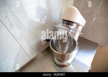 Weißer elektrischer Standmixer mit einem Rührschüssel aus Edelstahl und einem Drahtquirlaufsatz auf einer Arbeitsplatte aus Edelstahl in einem modernen Wohnbereich Stockfoto