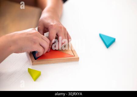 Die Hände eines Individuums stellen ein geometrisches Holzpuzzle mit bunten dreieckigen Teilen auf einer flachen weißen Oberfläche zusammen, wobei der Schwerpunkt auf der Platzierung einer Lasche liegt Stockfoto