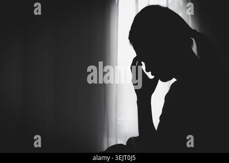 Silhouette einer einsamen jungen Frau, die sich depressiv und gestresst fühlt, Kopf in den Händen im dunklen Schlafzimmer sitzend, negative Emotionen und psychische Gesundheitskonzepte Stockfoto