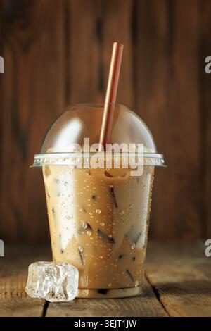 Eiskaffee in transparenter Plastikbecher zum Mitnehmen mit Eis und braunem Stroh, Kondenswasserperlen auf Holztisch Stockfoto
