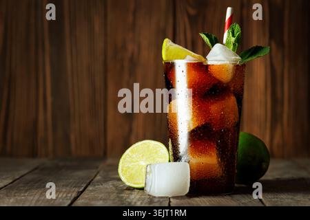 Cuba libre prickelnder Cola- und Rum-Cocktail ist im Glas mit Eis, Limettenscheiben und frischer Minze auf Holzoberfläche gekühlt Stockfoto