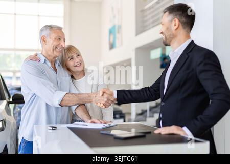 Glückliche ältere Ehepartner schütteln sich nach erfolgreichem Fahrzeugkauf die Hände mit dem Autoverkäufer Stockfoto
