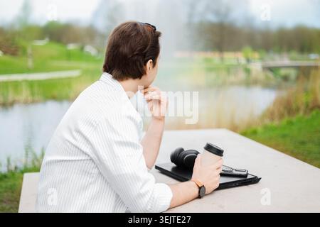 Rückansicht eines freischaffenden Mannes, der sich in einem Park im Freien erholt. Er macht eine Pause von der Laptop-Arbeit, hält einen Kaffee und denkt nach, während er die Natur betrachtet Stockfoto