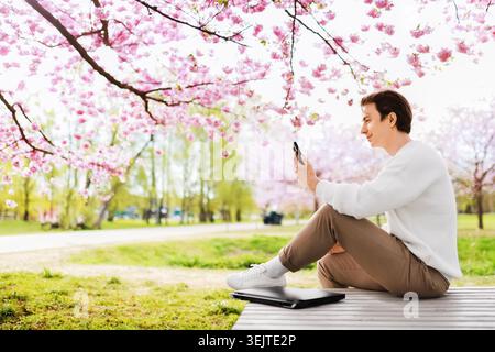 Glücklicher männlicher Freelancer, der eine Pause von der Fernarbeit im Freien macht. Junger Mann sitzt unter blühenden Kirschblüten mit einem Laptop und lächelt seinen Smart an Stockfoto