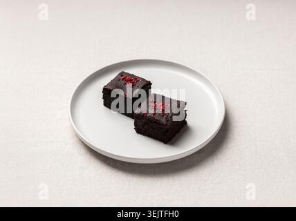 Zwei quadratische Stücke Schokoladenbrownie mit zerstoßenen Beeren auf einer hellen Keramikplatte. Weiche Beleuchtung und minimales Design machen ihn perfekt für Bak Stockfoto