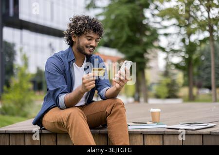 Junger Mann, der ein Handy und eine Kreditkarte für digitale Zahlungen oder Online-Einkäufe benutzt und draußen auf einer Holzbank sitzt, eine Kaffeetasse und Dokumente in der Nähe hat Stockfoto