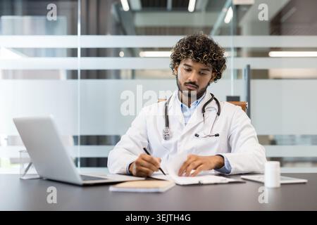 Junger männlicher Arzt, der Patienteninformationen analysiert und auf eine Zwischenablage schreibt, während er an einem Schreibtisch mit Laptop und Stethoskop sitzt, in einer modernen medizinischen Praxis Stockfoto