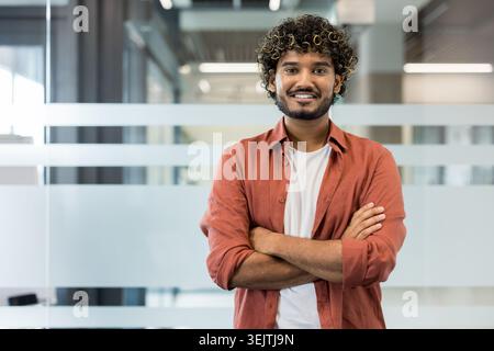 Junger, professioneller Mann mit lockigen Haaren und Bart, der in einem modernen Büro steht und selbstbewusst lächelt und die Arme überkreuzt. Er steht für eine freundliche und zugängliche Geschäftspräsenz Stockfoto
