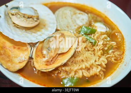 Eine lebendige Nahaufnahme scharfer koreanischer Meeresfrüchte-Ramen in einer weißen Schüssel mit lockigen Nudeln, kräftiger roter Brühe, großen offenen Venusmuscheln und grünen Zwiebeln. Stockfoto