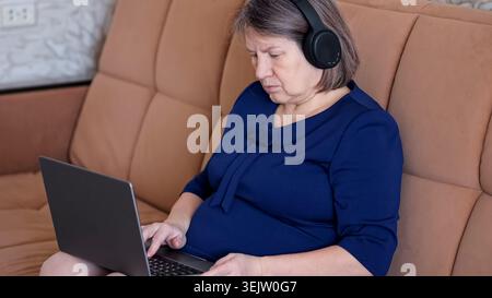Ältere Frau, die Kopfhörer trägt, während sie sich auf einen Laptop konzentriert, bequem auf einer Couch sitzt und zu Hause Online-Lernen oder Fernarbeit absolviert Stockfoto