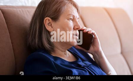 Ältere Frau zu Hause, die auf einem Sofa sitzt, ein Mobiltelefon am Ohr hält und mit besorgtem Ausdruck spricht, wichtige Nachrichten übermittelt oder Stockfoto