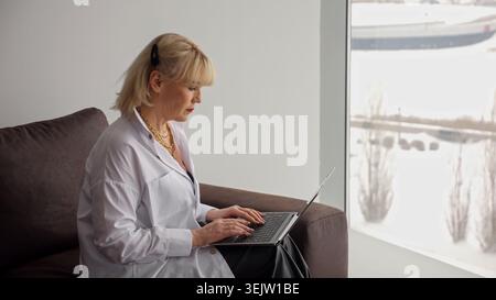 Blonde Frau, die auf einem Laptop schreibt, an Fernarbeit oder Online-Bildung teilnimmt und dabei bequem auf einer Couch am Fenster sitzt und eine Winterlandschaft bietet Stockfoto