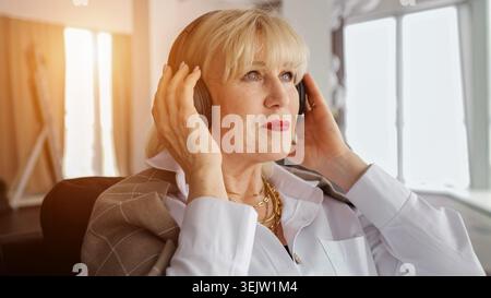 Blonde Seniorin sitzt drinnen mit schnurlosen Kopfhörern, genießt Musik und Entspannung zu Hause, friedlich und zufrieden während der Freizeit und Stockfoto